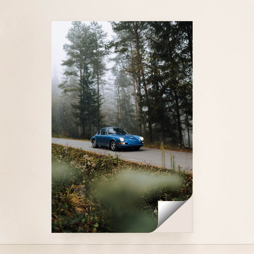 This photo shows a classic blue Porsche driving along a misty forest road with wildflowers in the foreground, minimalist unframed style.