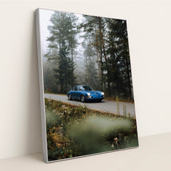 This photo shows a classic blue Porsche driving along a misty forest road with wildflowers in the foreground, in a silver frame.