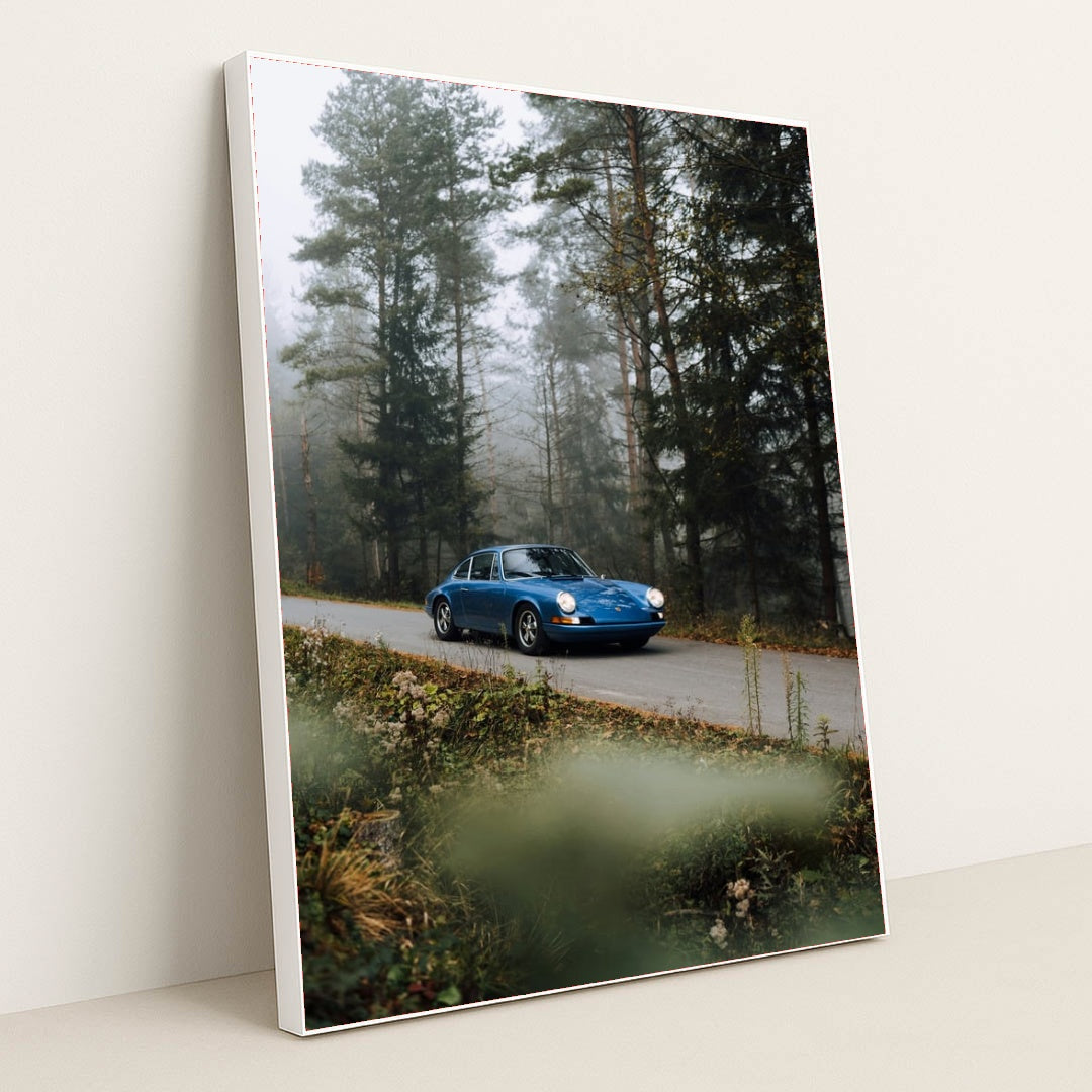 This photo shows a classic blue Porsche driving along a misty forest road with wildflowers in the foreground, in a white frame.