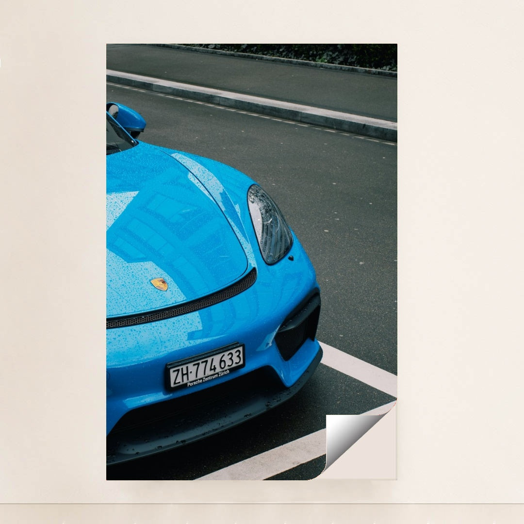 This photo shows the front of a blue Porsche sports car with raindrops, parked on wet pavement, unframed.