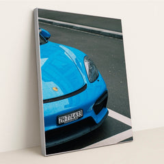 This photo shows the front of a blue Porsche sports car with raindrops, parked on wet pavement, in a silver frame.