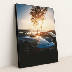This photo shows a vintage light blue Porsche 911 parked at sunset with a tree and mountains in the background, in a black frame.