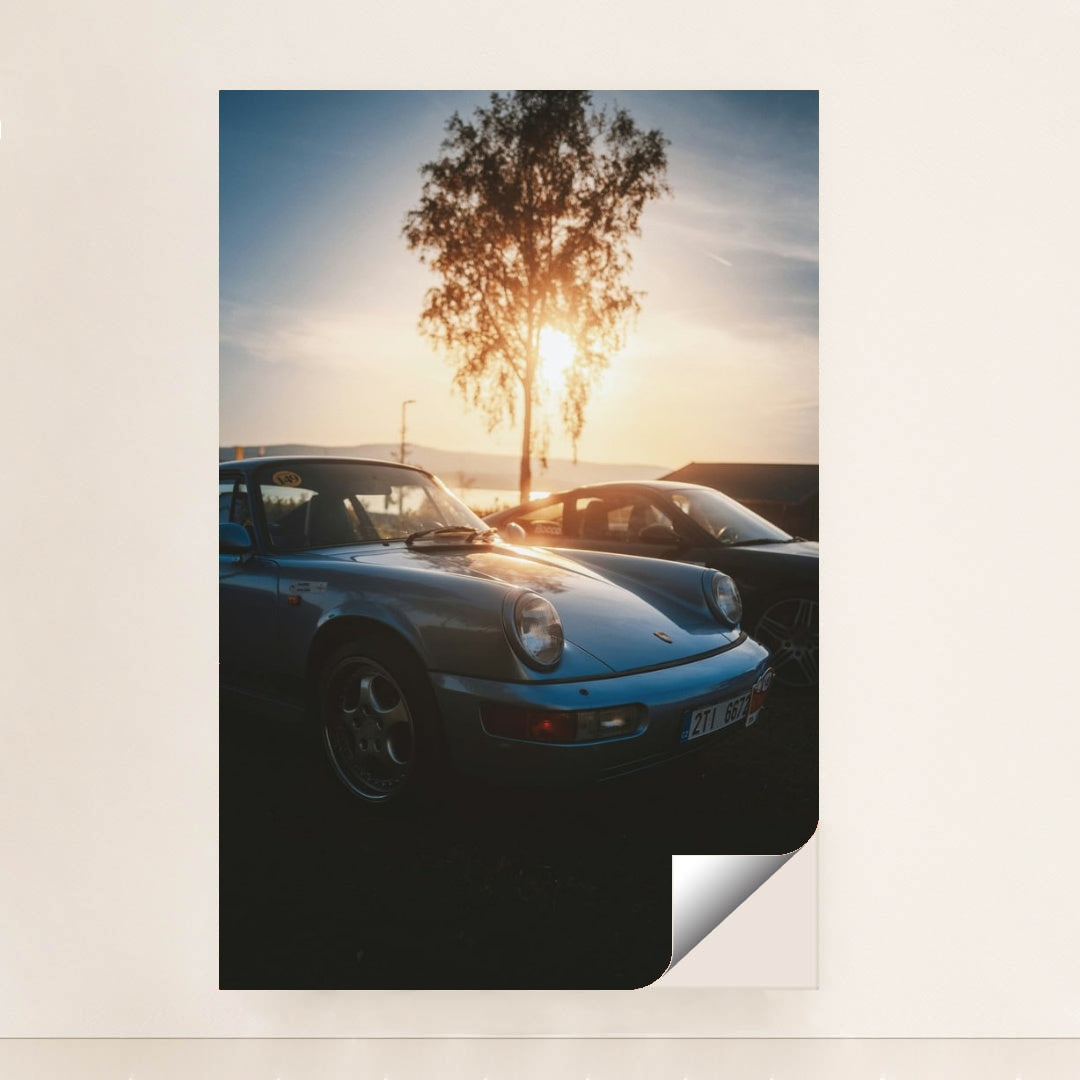 This photo shows a vintage light blue Porsche 911 parked at sunset with a tree and mountains in the background, minimalist unframed style.
