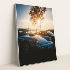 This photo shows a vintage light blue Porsche 911 parked at sunset with a tree and mountains in the background, in a silver frame.