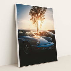 This photo shows a vintage light blue Porsche 911 parked at sunset with a tree and mountains in the background, in a white frame.