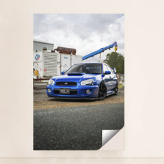 This photo shows a blue Subaru WRX with black wheels parked in an industrial lot, unframed style.