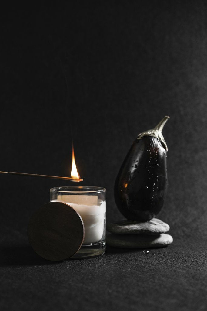 Mikhail Nilov - A minimalist still life featuring a lit candle, an eggplant, and stones on a dark surface.