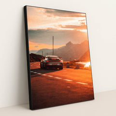 This photo shows a silver Porsche sports car driving on a winding mountain road at sunset, in a black frame.