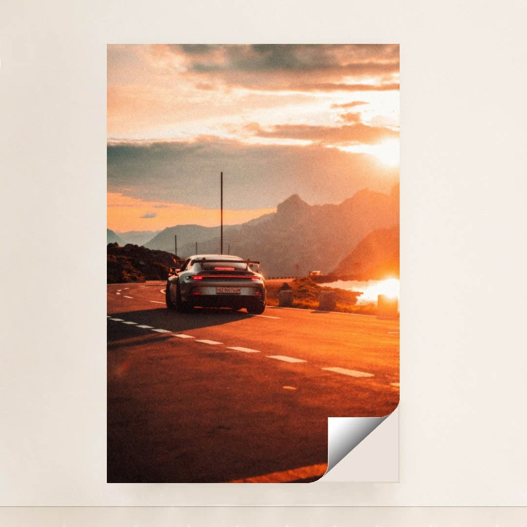 This photo shows a silver Porsche sports car driving on a winding mountain road at sunset, minimalist unframed style.