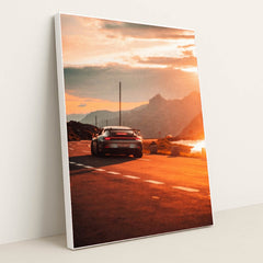 This photo shows a silver Porsche sports car driving on a winding mountain road at sunset, in a white frame.
