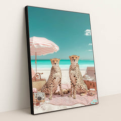 This artwork shows two cheetahs sitting on a pink picnic blanket under a striped umbrella at a bright beach, in a black frame.