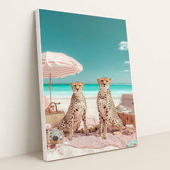 This artwork shows two cheetahs sitting on a pink picnic blanket under a striped umbrella at a bright beach, in a white frame.