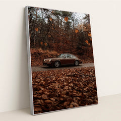 This photo shows a brown classic Porsche 911 coupe parked on a road surrounded by autumn leaves in a forest, in a silver frame.