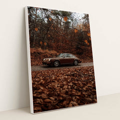 This photo shows a brown classic Porsche 911 coupe parked on a road surrounded by autumn leaves in a forest, in a white frame.