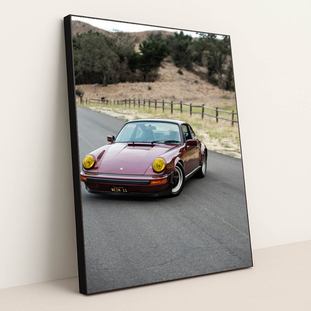 This photo shows a vintage maroon Porsche 911 with yellow headlights driving along a country road, in a black frame.