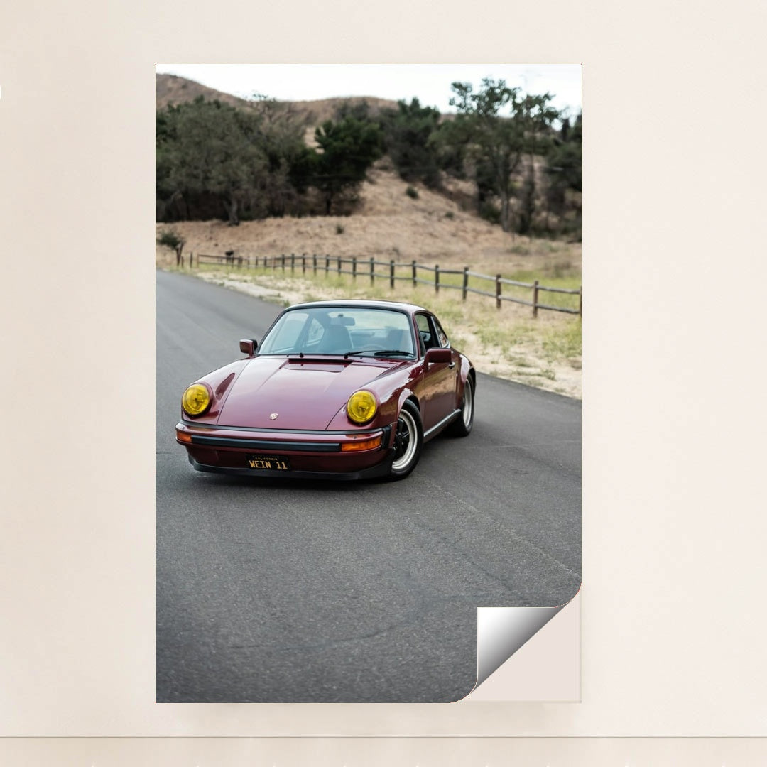 This photo shows a vintage maroon Porsche 911 with yellow headlights driving along a country road, unframed style.