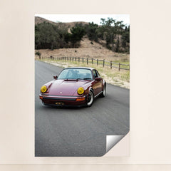 This photo shows a vintage maroon Porsche 911 with yellow headlights driving along a country road, unframed style.