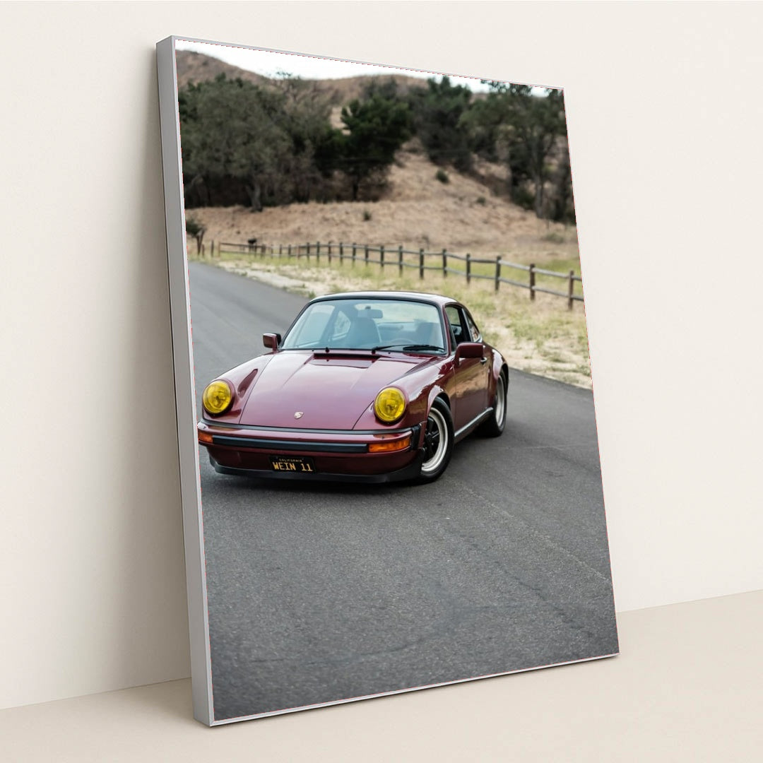 This photo shows a vintage maroon Porsche 911 with yellow headlights driving along a country road, in a silver frame.