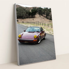 This photo shows a vintage maroon Porsche 911 with yellow headlights driving along a country road, in a silver frame.