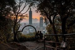 Jimmy Liao - A couple sits on a scenic hill, admiring Taipei 101 at sunset, framed by lush trees.