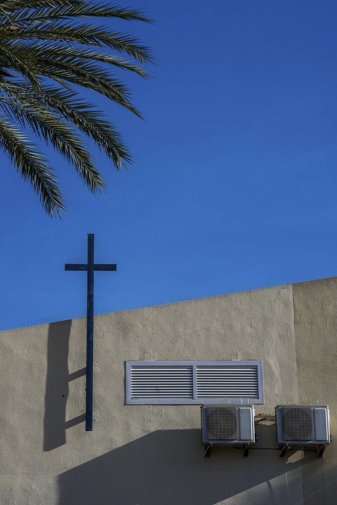 Victor Moragriega - A modern church facade with a cross, palm tree, and air conditioners against a vivid blue sky.