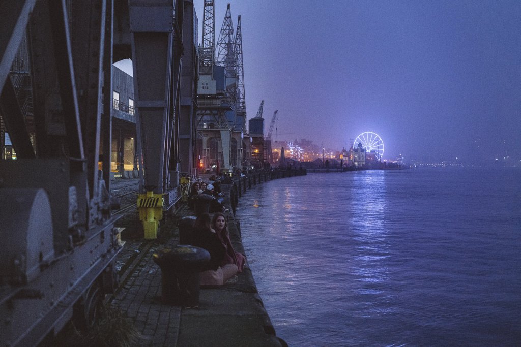 Ricardo Oliveira - Dusk at a bustling waterfront with a glowing Ferris wheel and shipyard ambiance.