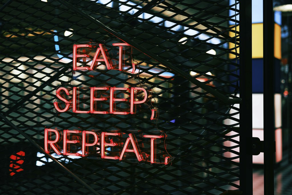 Yusuf Timur Çelik - Close-up of a vibrant neon sign with the slogan 'Eat, Sleep, Repeat' on a metal grate background.
