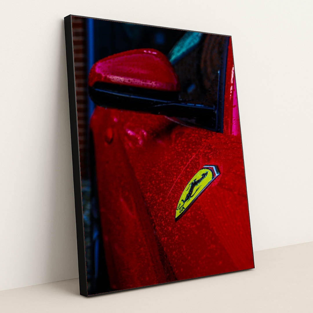 This photo shows the close-up of a red Ferrari’s fender and badge covered in rain droplets, in a black frame.