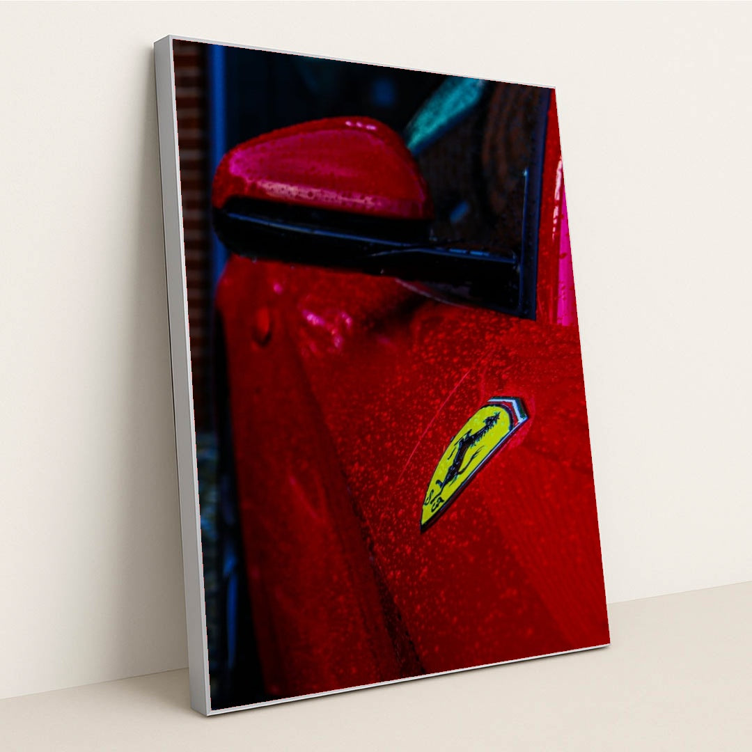This photo shows the close-up of a red Ferrari’s fender and badge covered in rain droplets, in a silver frame.