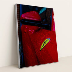 This photo shows the close-up of a red Ferrari’s fender and badge covered in rain droplets, in a silver frame.