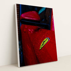 This photo shows the close-up of a red Ferrari’s fender and badge covered in rain droplets, in a white frame.