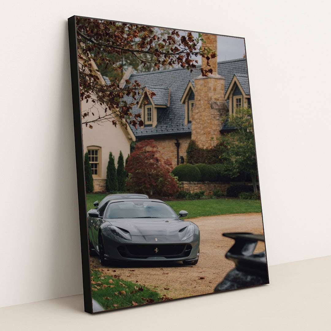 This photo shows a grey Ferrari parked on a stone driveway in front of a large estate with autumn foliage, in a black frame.