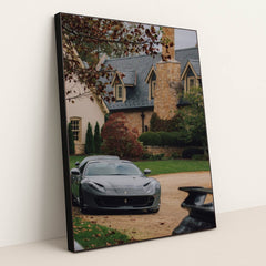 This photo shows a grey Ferrari parked on a stone driveway in front of a large estate with autumn foliage, in a black frame.