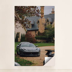 This photo shows a grey Ferrari parked on a stone driveway in front of a large estate with autumn foliage, minimalist unframed style.