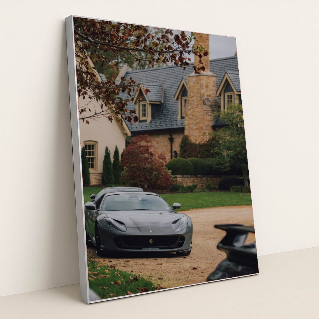 This photo shows a grey Ferrari parked on a stone driveway in front of a large estate with autumn foliage, in a silver frame.