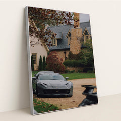 This photo shows a grey Ferrari parked on a stone driveway in front of a large estate with autumn foliage, in a silver frame.