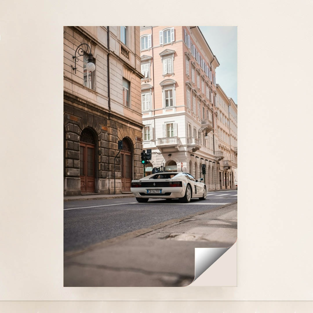 This photo shows a white Ferrari Testarossa driving through a European city street lined with historic pastel buildings, minimalist unframed style.