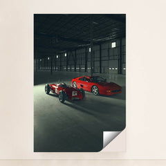 This photo shows a vintage red Ferrari race car and a red Ferrari coupe parked inside a large industrial warehouse, minimalist unframed style.