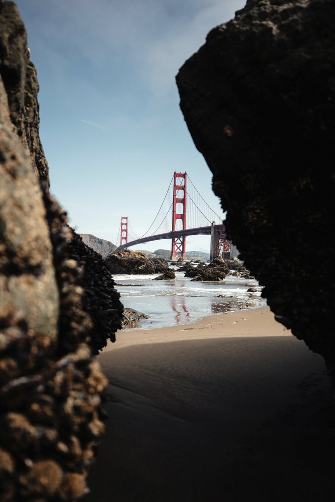 Dominique Barrera - Iconic view of the Golden Gate Bridge through coastal rocks on a clear day.