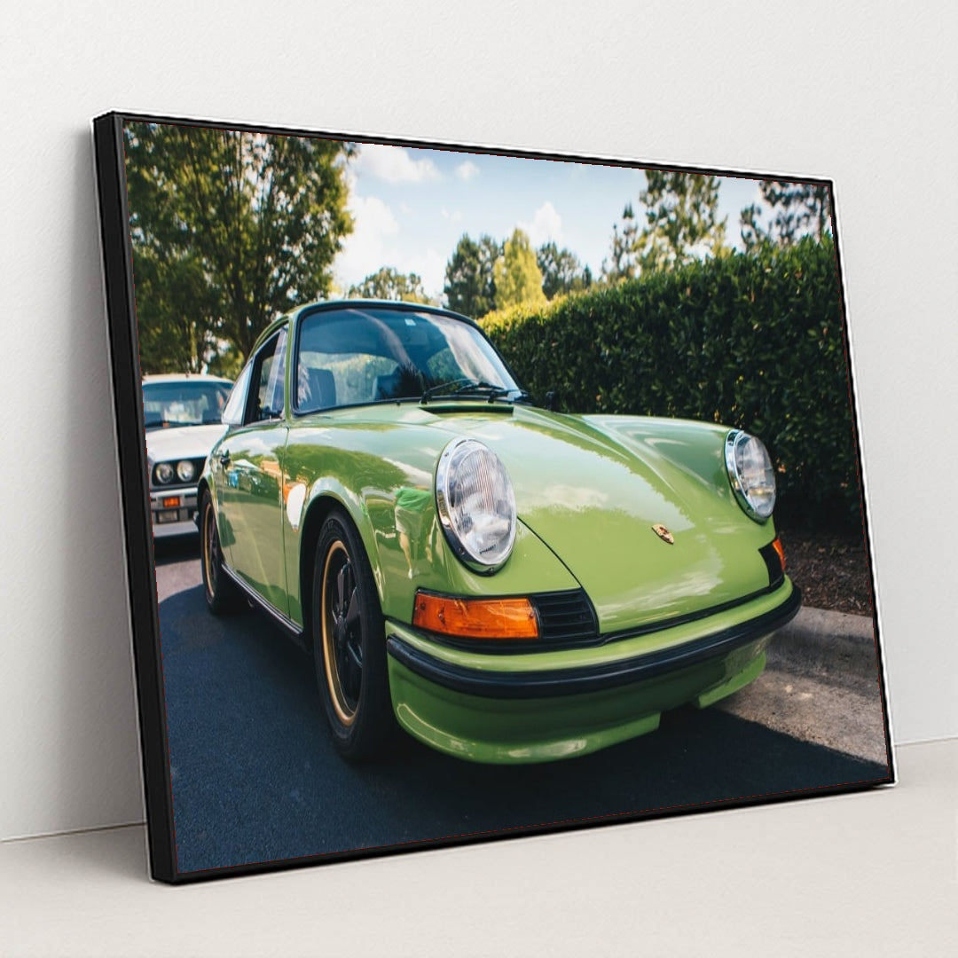 This photo shows a classic Porsche 911 in light green parked on a sunny street with trees in the background, in a black frame.