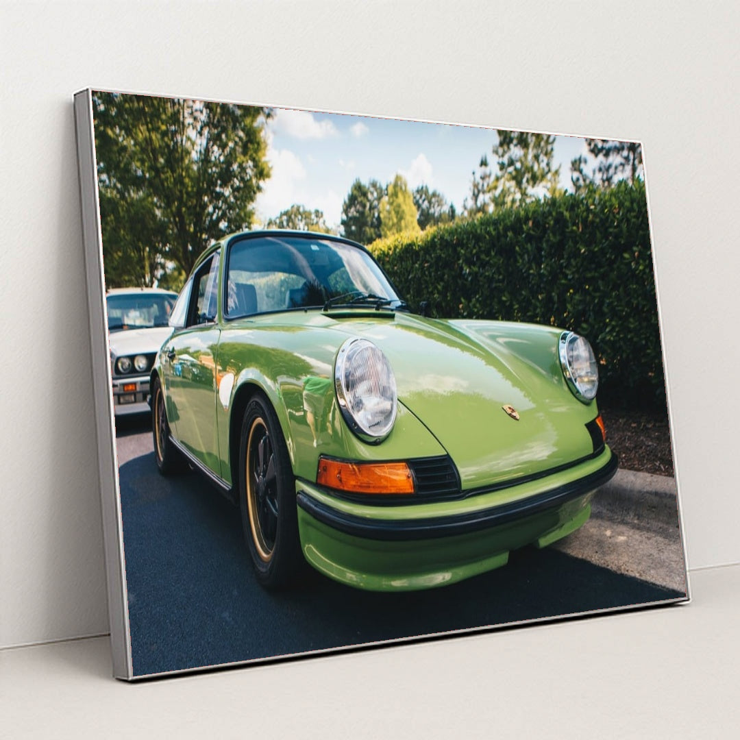 This photo shows a classic Porsche 911 in light green parked on a sunny street with trees in the background, in a silver frame.