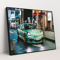 This photo shows a mint green Porsche sports car with gold wheels driving through a vibrant city street at night, in a black frame.