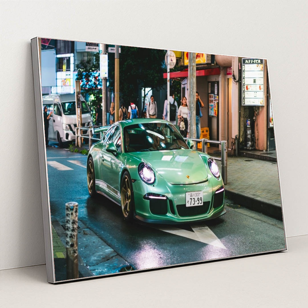 This photo shows a mint green Porsche sports car with gold wheels driving through a vibrant city street at night, in a silver frame.