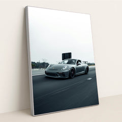 This photo shows a gray Porsche sports car driving fast on a highway with a blurred background, in a silver frame.