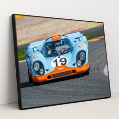 This photo shows a vintage blue and orange Porsche race car with the number 19 rounding a track corner, in a black frame.