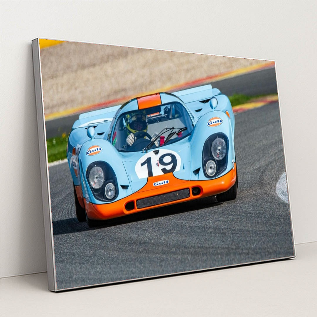 This photo shows a vintage blue and orange Porsche race car with the number 19 rounding a track corner, in a silver frame.