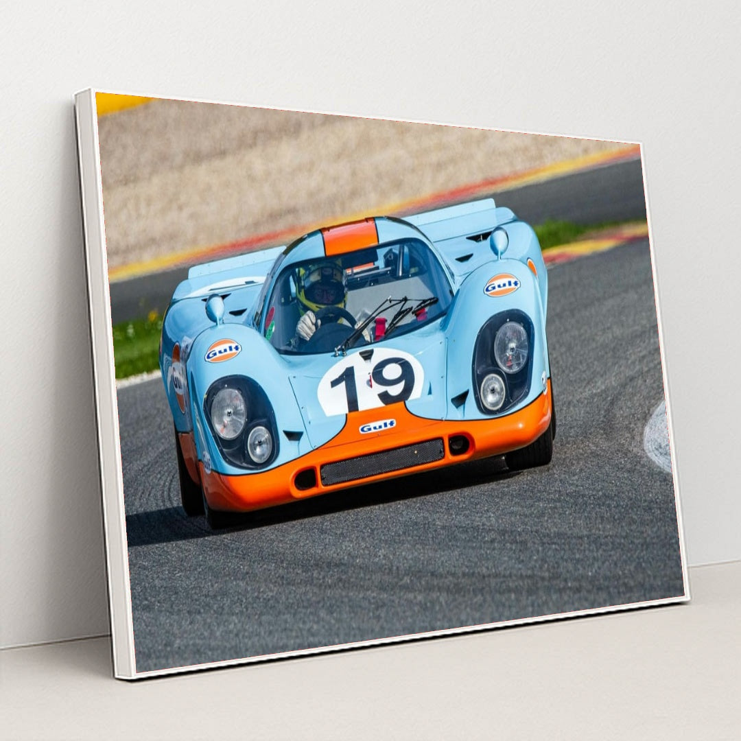This photo shows a vintage blue and orange Porsche race car with the number 19 rounding a track corner, in a white frame.
