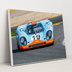 This photo shows a vintage blue and orange Porsche race car with the number 19 rounding a track corner, in a white frame.