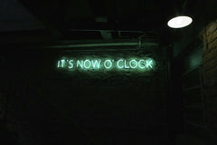 Freitas Junior - Green neon sign in a dimly lit room showcasing 'It's Now O' Clock'.
