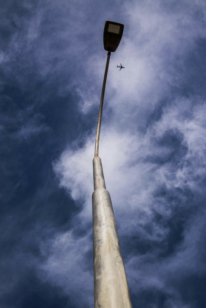 Victor Moragriega - Low-angle shot of a streetlight and airplane against a cloudy blue sky.
